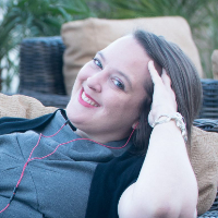 A woman with long brown hair smiles while reclining on an outdoor wicker chair, wearing a gray top, black cardigan, and red earphones. [gen-ai]