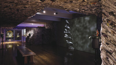 An art gallery with walls and ceiling covered in crumpled paper, wooden floors, benches, and a projected image of a plant on one wall.