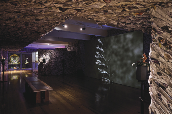 An art gallery with walls and ceiling covered in crumpled paper, wooden floors, benches, and a projected image of a plant on one wall.