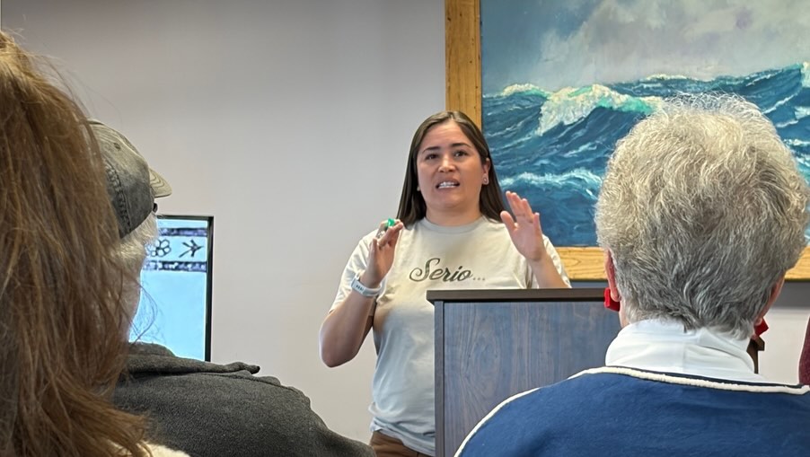 A woman stands behind a podium speaking to an audience, with a large ocean painting and a screen in the background.