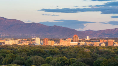 A city skyline with mid-rise buildings is set against a backdrop of mountains under a partly cloudy sky, with trees in the foreground.