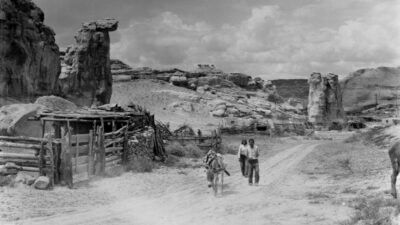 Two people walk beside a donkey on a dirt road near a wooden fence and rocky landscape under a cloudy sky.