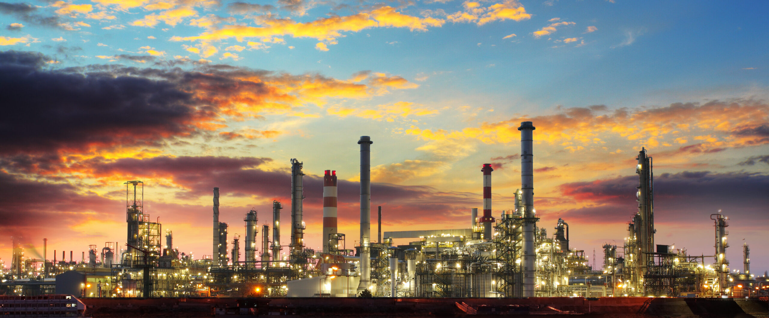 An oil refinery with multiple towers and industrial structures is seen against a colorful sunset sky.