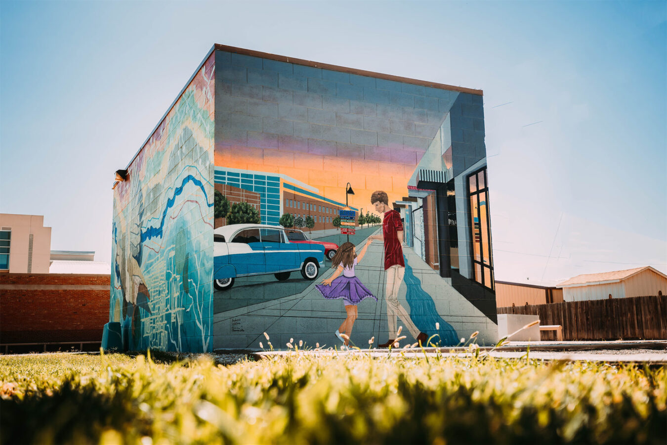 A small building with a colorful mural depicting a man and girl dancing in front of a blue vintage car, set in an urban and grassy area under a clear sky.