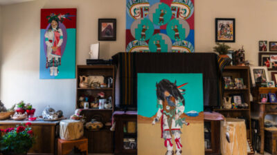 Art-filled living room with paintings, shelves of figurines, framed photos, potted plants, and a drum arranged against a white wall.