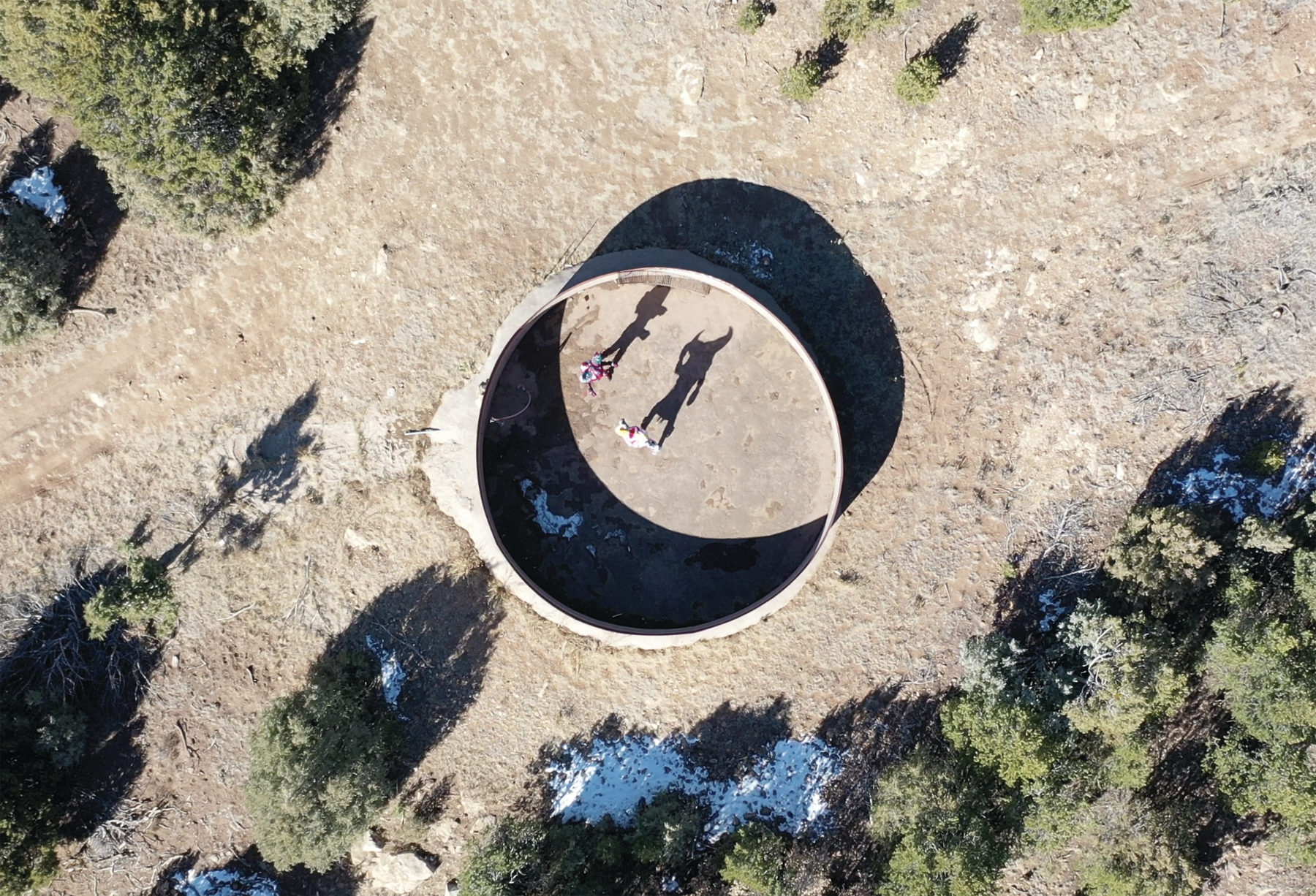 Aerial view of two people standing inside a large, circular concrete structure casting shadows, surrounded by dry terrain and sparse vegetation.