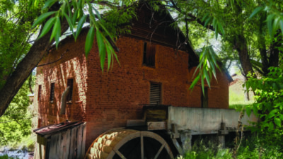 A brick watermill with a large wooden wheel is surrounded by green trees and foliage, with sunlight filtering through the leaves.