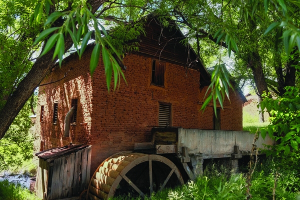 A brick watermill with a large wooden wheel is surrounded by green trees and foliage, with sunlight filtering through the leaves.