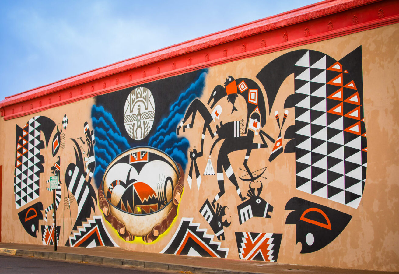 Colorful mural with geometric and symbolic Native American patterns, including animals, people, and a central bowl motif, painted on an exterior wall under a red roof.