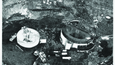 A black and white aerial view of a construction site with circular structures, scattered materials, vehicles, and uneven terrain.