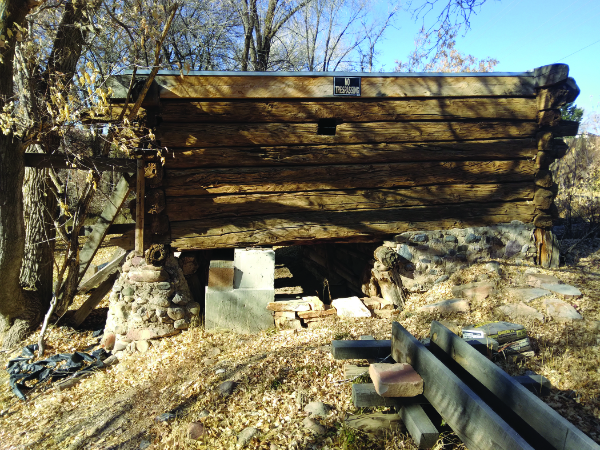 A weathered wooden structure with log walls stands elevated on stone supports, surrounded by dry leaves, fallen branches, and scattered debris.