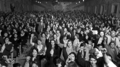 A large crowd of people, mostly women, sit closely together indoors, raising their fists in unison, suggesting a protest or rally.