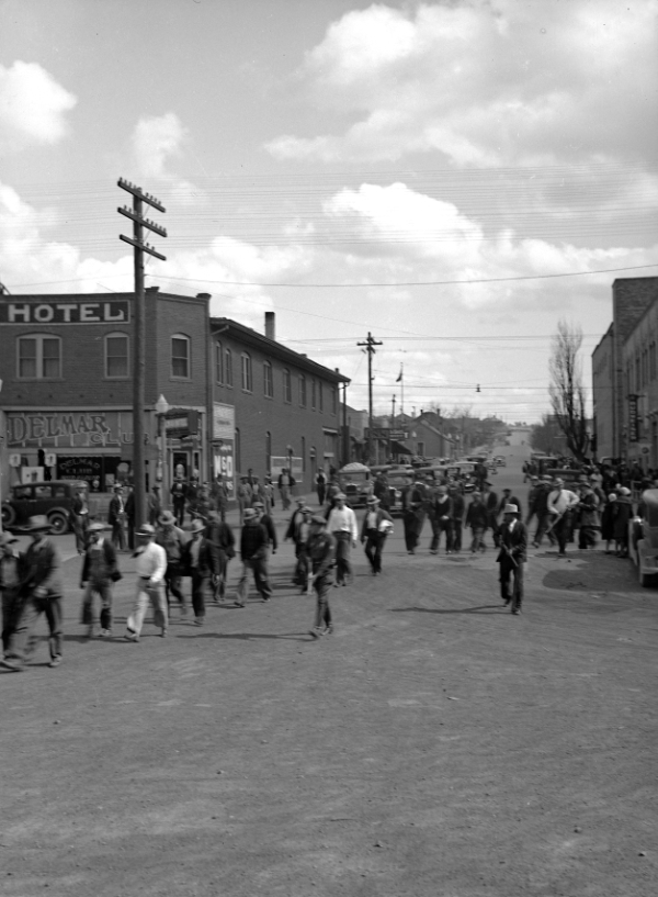 Black and white photo of people walking on a busy street in front of buildings, including a hotel and the Delmar, with cars parked along the road.