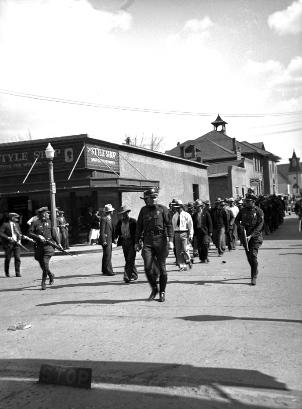 Police officers and a group of people walk down a city street, passing by storefronts and buildings under a partly cloudy sky.