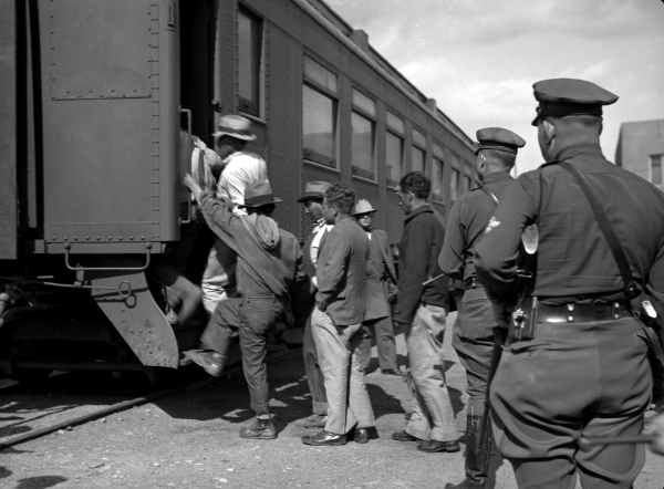 A group of people board a train car while uniformed officers stand nearby, observing.