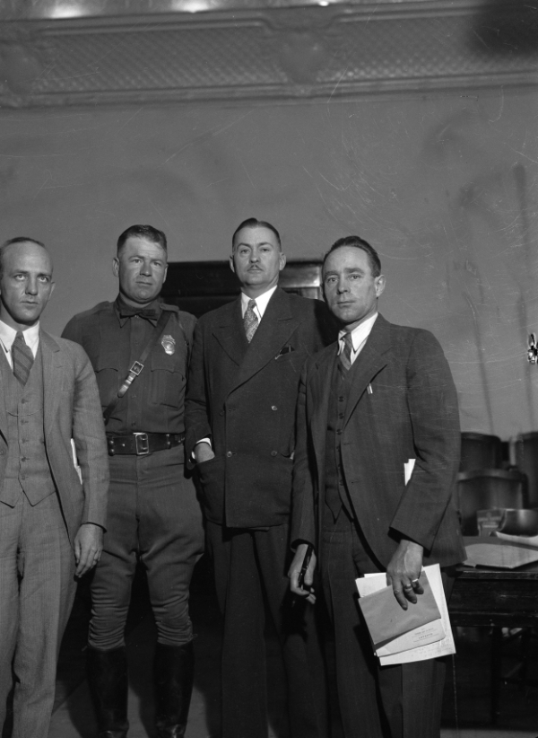 Four men in suits, one wearing a police uniform, stand indoors next to each other; two are holding papers.