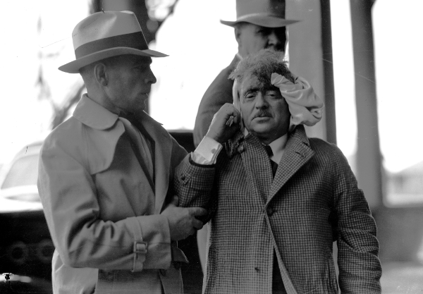 Two men stand outside; one, in a coat and hat, presses a cloth to the head of the other man, who appears injured and is being assisted.