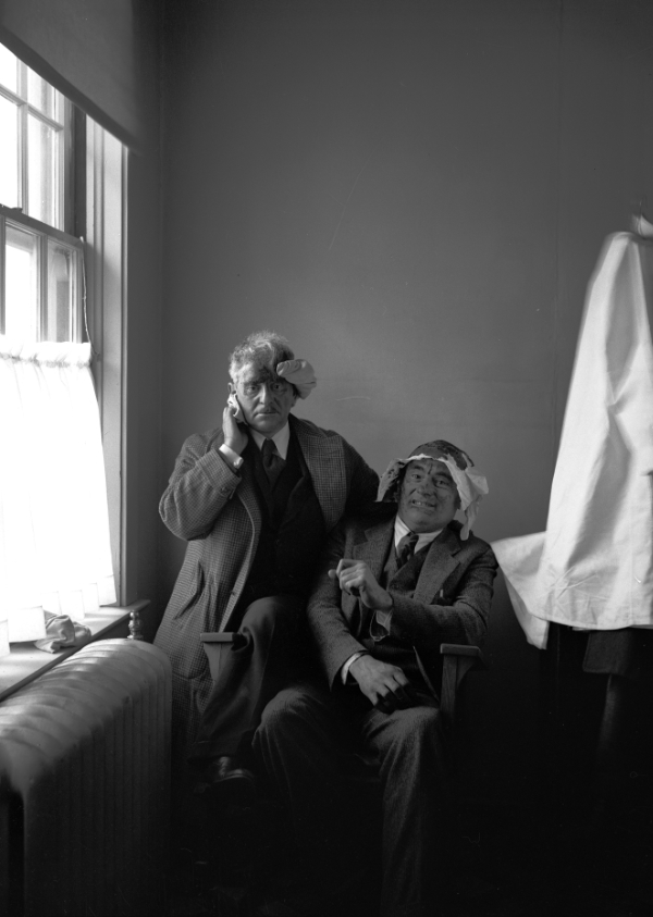 Two men sit by a window, each holding a cloth to their heads as if nursing injuries. One man is seated, while the other stands behind him. Both wear suits and serious expressions.