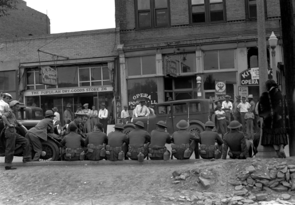 A row of uniformed men sit on the curb facing a group of people standing in front of storefronts on a city street; cars and a woman walking are also visible.