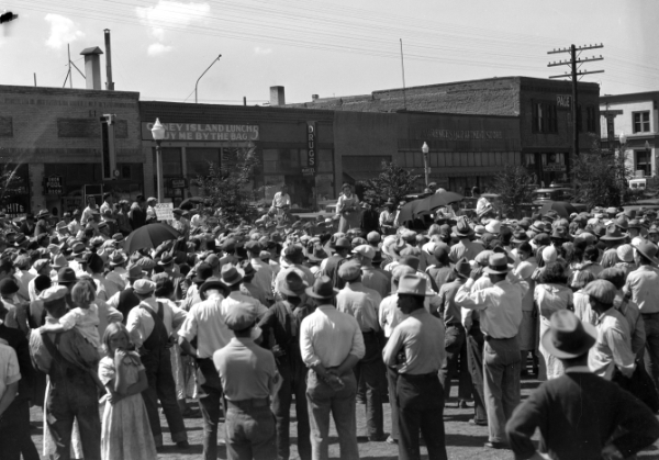 A large crowd of people, mostly wearing hats, gathers outdoors in front of buildings to listen to a speaker during the daytime.