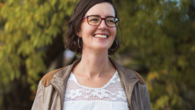 Woman with short brown hair and glasses, wearing a light jacket and white top, standing outdoors in front of leafy green trees, smiling.
