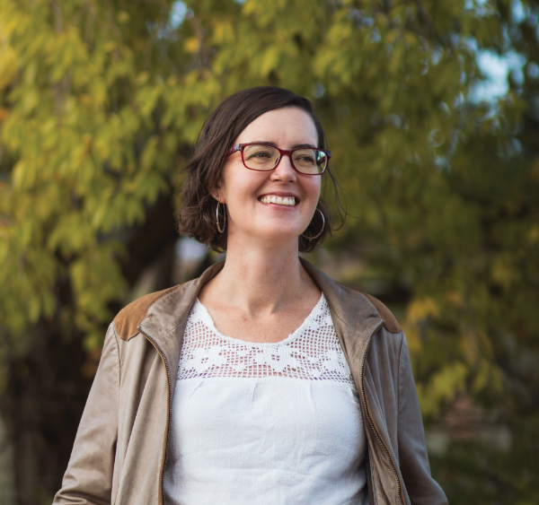 Woman with short brown hair and glasses, wearing a light jacket and white top, standing outdoors in front of leafy green trees, smiling.