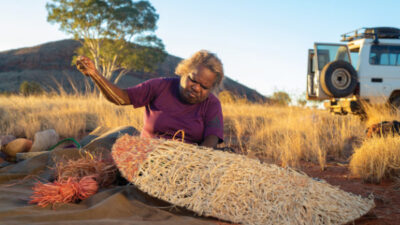 An older woman sits outdoors on red soil, weaving a large basket from natural fibers, with dry grass, a tree, and a vehicle in the background.