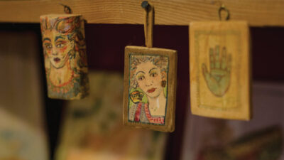 Three small painted wooden plaques hang from a shelf, featuring two illustrated faces and one palmistry hand design, with various blurred objects in the background.