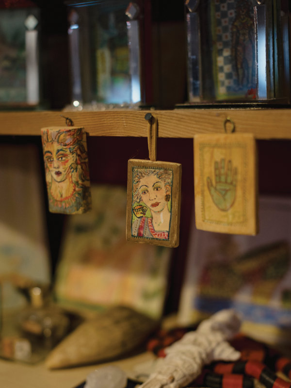 Three small painted wooden plaques hang from a shelf, featuring two illustrated faces and one palmistry hand design, with various blurred objects in the background.