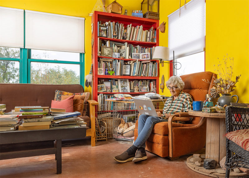 An older person sits in an orange armchair using a laptop in a bright, yellow-walled room with bookshelves, a couch, and stacked books.