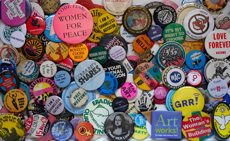 A collection of colorful pins and buttons with various slogans, symbols, and messages related to activism, art, and social causes, arranged in a dense overlapping pattern.