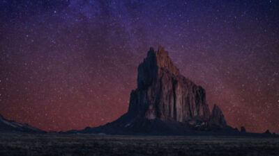 A large, jagged rock formation rises from a flat desert landscape under a purple and red star-filled night sky.