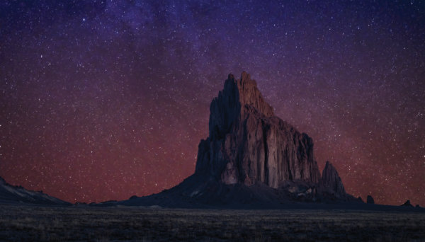 A large, jagged rock formation rises from a flat desert landscape under a purple and red star-filled night sky.