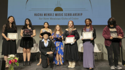 Seven young women and one adult pose on stage, some holding certificates, in front of a screen displaying Nacha Mendez Music Scholarship for New Mexican Girls of Color.