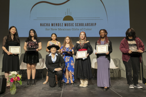 Seven young women and one adult pose on stage, some holding certificates, in front of a screen displaying Nacha Mendez Music Scholarship for New Mexican Girls of Color.