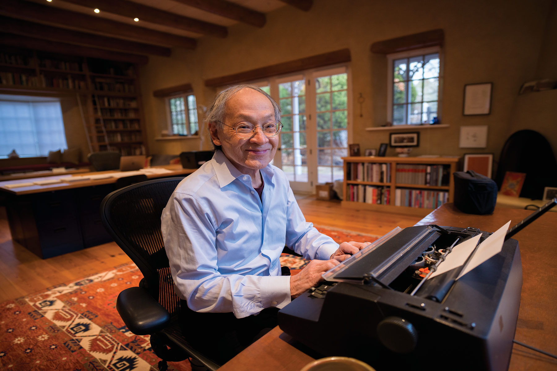 An older man in a light blue shirt sits at a desk, typing on a typewriter in a spacious, book-filled home office.