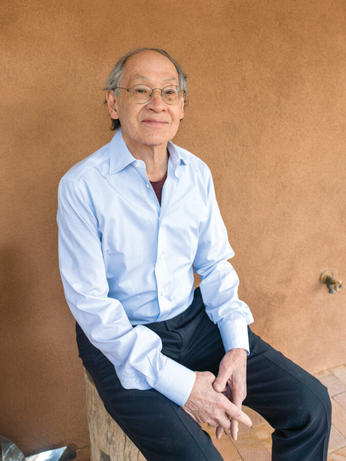 An older man with glasses, wearing a light blue shirt and dark pants, sits on a wooden seat against a tan stucco wall.