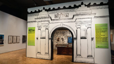 Exhibit entrance designed as a black-and-white illustrated archway with green information panels; artwork and objects displayed inside and on surrounding walls.