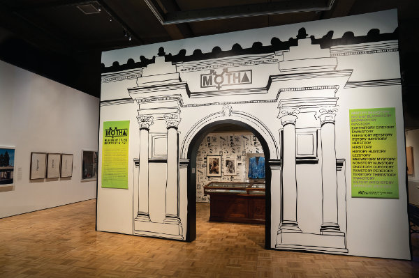 Exhibit entrance designed as a black-and-white illustrated archway with green information panels; artwork and objects displayed inside and on surrounding walls.