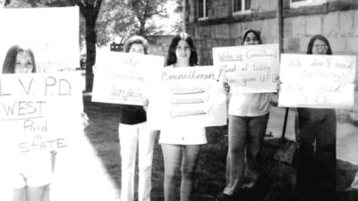 Five people stand outside holding protest signs with messages about unfair wages and cost of living increases.