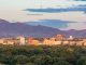 City skyline with several tall buildings set against a backdrop of tree-filled areas and distant mountains under a partly cloudy sky at sunset.