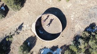 Aerial view of a circular concrete structure with two people and a bicycle inside, casting long shadows. The structure is surrounded by dirt, patches of vegetation, and small trees.