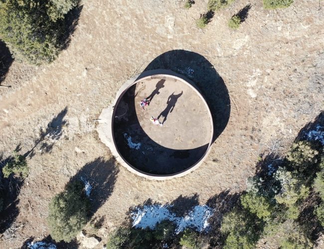 Aerial view of a circular concrete structure with two people and a bicycle inside, casting long shadows. The structure is surrounded by dirt, patches of vegetation, and small trees.