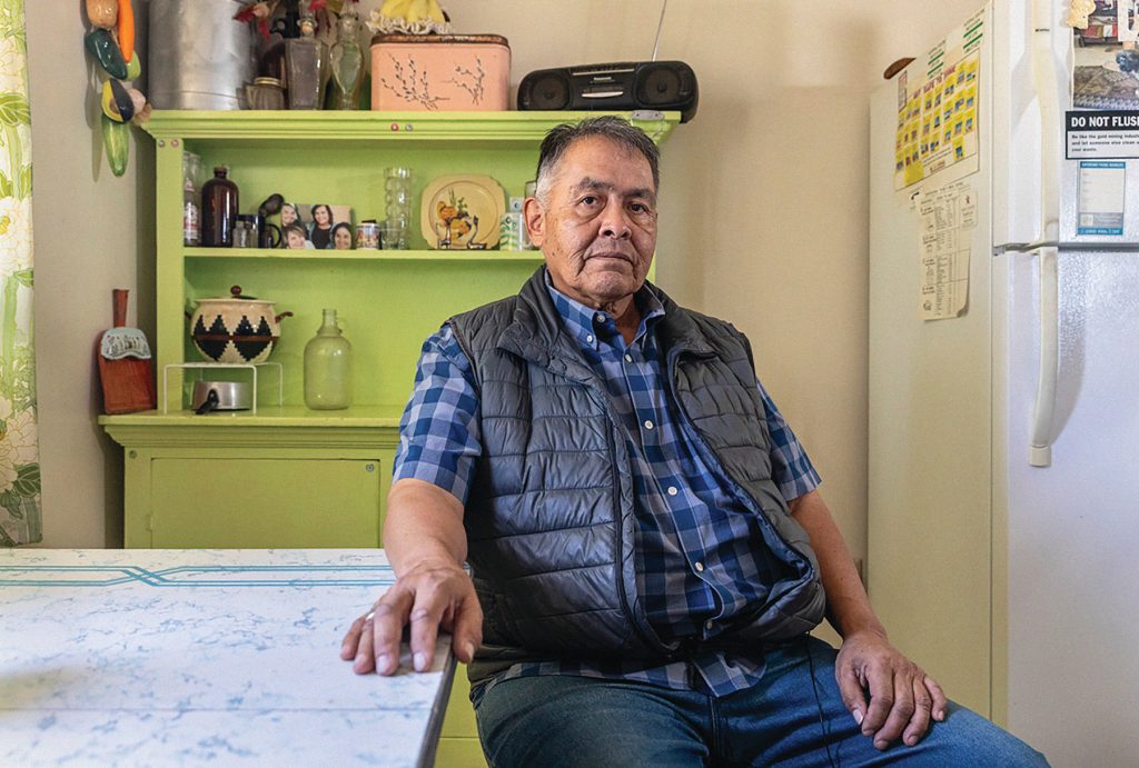 An older man wearing a plaid shirt and gray vest sits at a kitchen table. Behind him is a green hutch with family photos and decorative items. A refrigerator and a wall calendar are visible on the right.