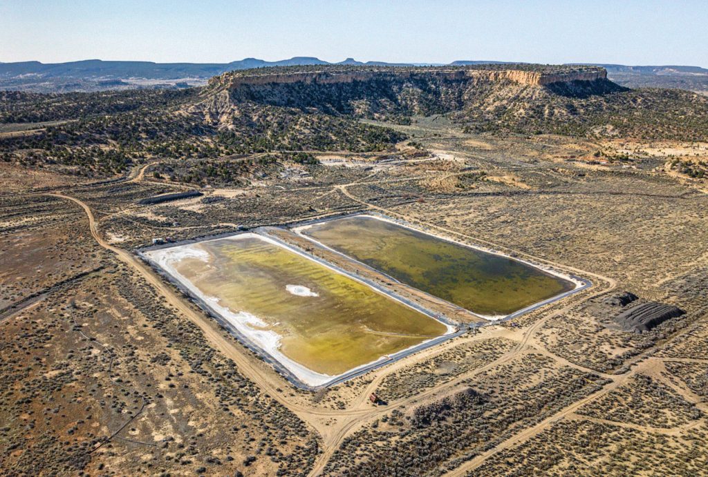 Aerial view of two large, rectangular evaporation ponds with yellow-green water in a dry, desert landscape, surrounded by dirt roads and low vegetation, with mesas in the background.