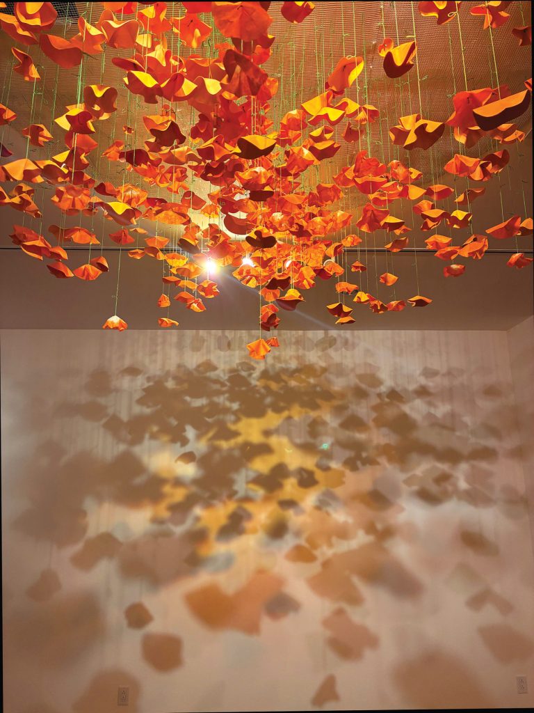 Orange and red paper sculptures hang from the ceiling on strings, illuminated by a central light that casts intricate shadows of the shapes onto the wall below.