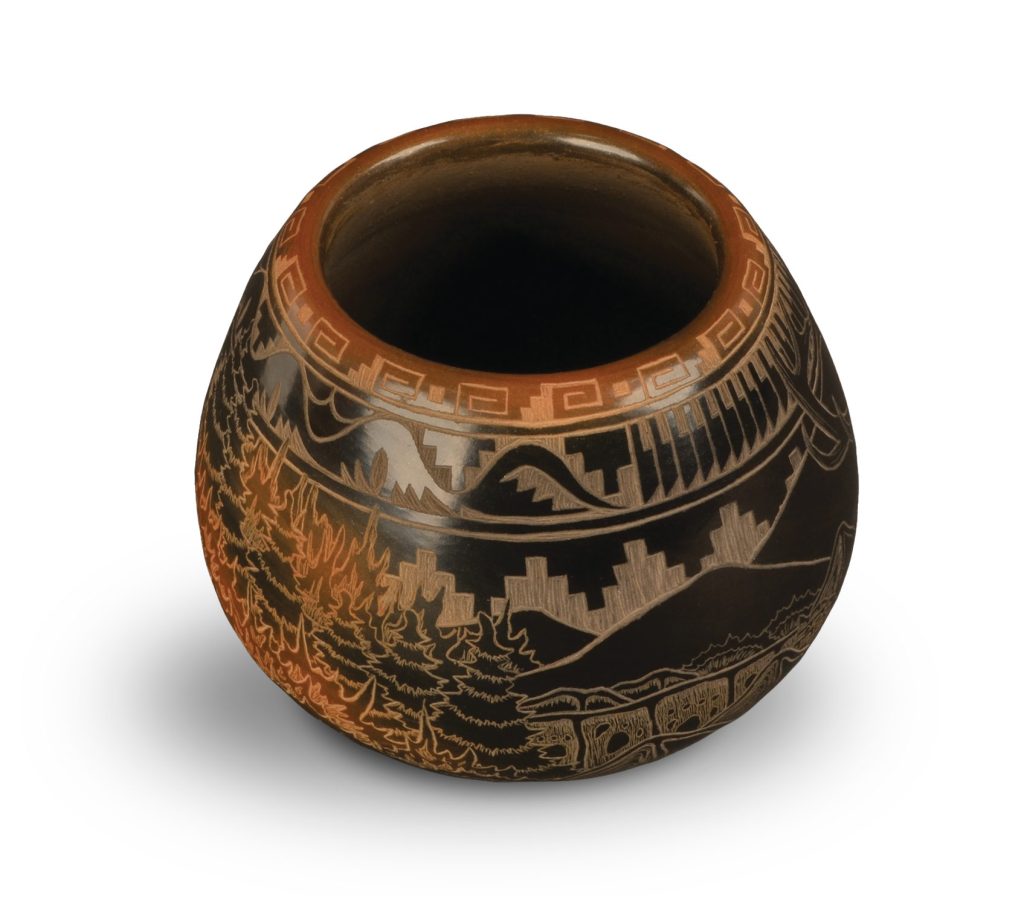 A round, handcrafted pottery vase with intricate etched designs, including geometric patterns near the rim and detailed scenes of buildings and trees around the body, on a white background.