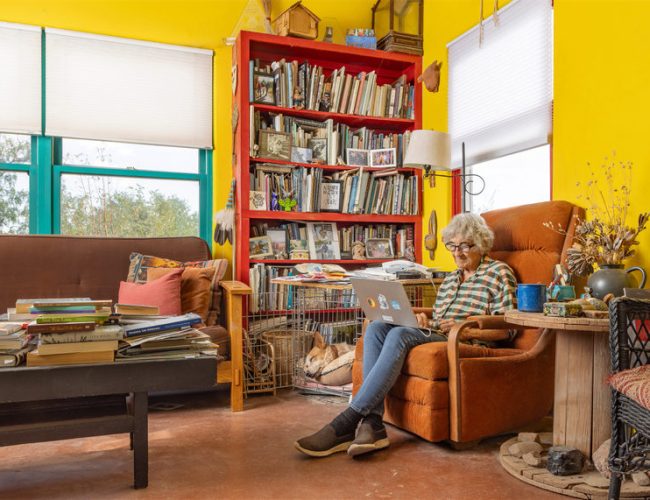 An older person with gray hair sits in an orange armchair using a laptop in a colorful living room with yellow walls, a bookshelf full of books, a couch, a coffee table with stacks of books, and a dog lying in a crate.