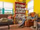 An older person with gray hair sits in an orange armchair using a laptop in a colorful living room with yellow walls, a bookshelf full of books, a couch, a coffee table with stacks of books, and a dog lying in a crate.