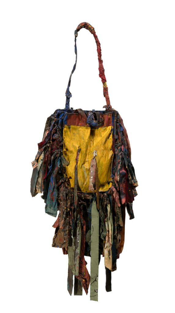 A colorful, ragged shoulder bag made of layered, tattered fabric strips in various colors, with a prominent yellow pocket on the front and a handle wrapped in fabric, set against a white background.